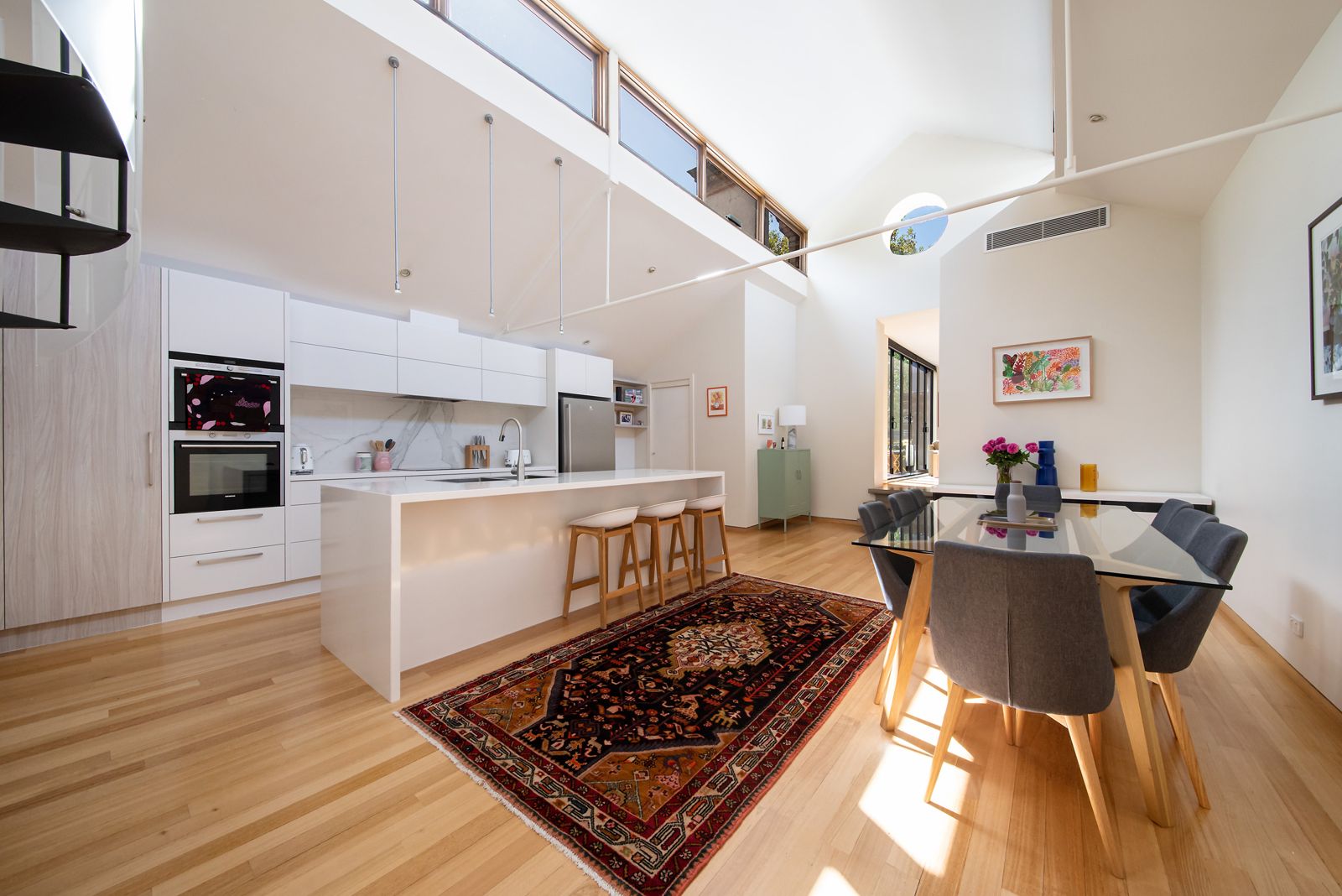 Open-plan kitchen and dining beneath soaring ceilings and clerestory windows, with the black steel spiral stair rising to the mezzanine at 2/1A Stanley Street, Collingwood